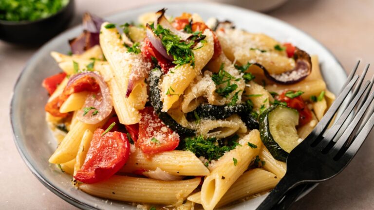 A plate of penne pasta mixed with roasted vegetables, including zucchini, tomatoes, and red onions, topped with grated cheese and chopped parsley. A fork rests on the plate.