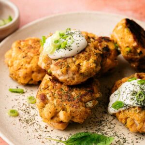 A plate of golden-brown Salmon Patties topped with sour cream, chopped green onions, and dill, served on a beige plate and garnished with fresh basil leaves.