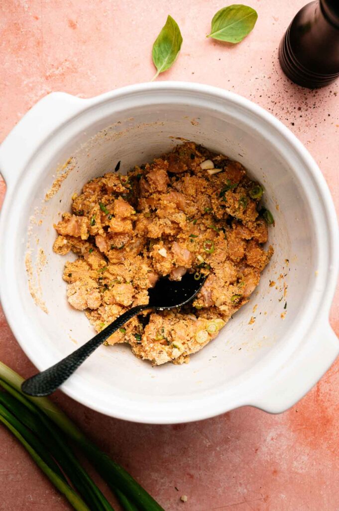 A white bowl with a mixture of ground meat, spices, and herbs—perfect for forming delicious salmon patties—with a black spoon inside. Green onions and basil leaves are nearby on a pink surface.