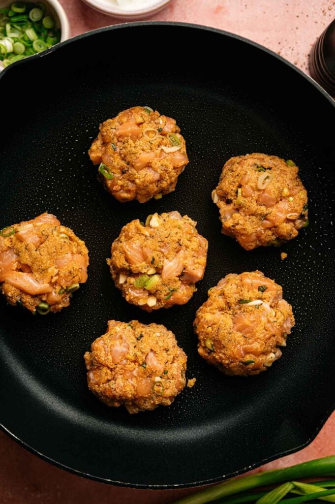 Five raw salmon patties with green onions and seasonings are arranged in a black skillet, ready to be cooked. These flavorful salmon patties promise a delicious meal packed with freshness and vibrant taste.