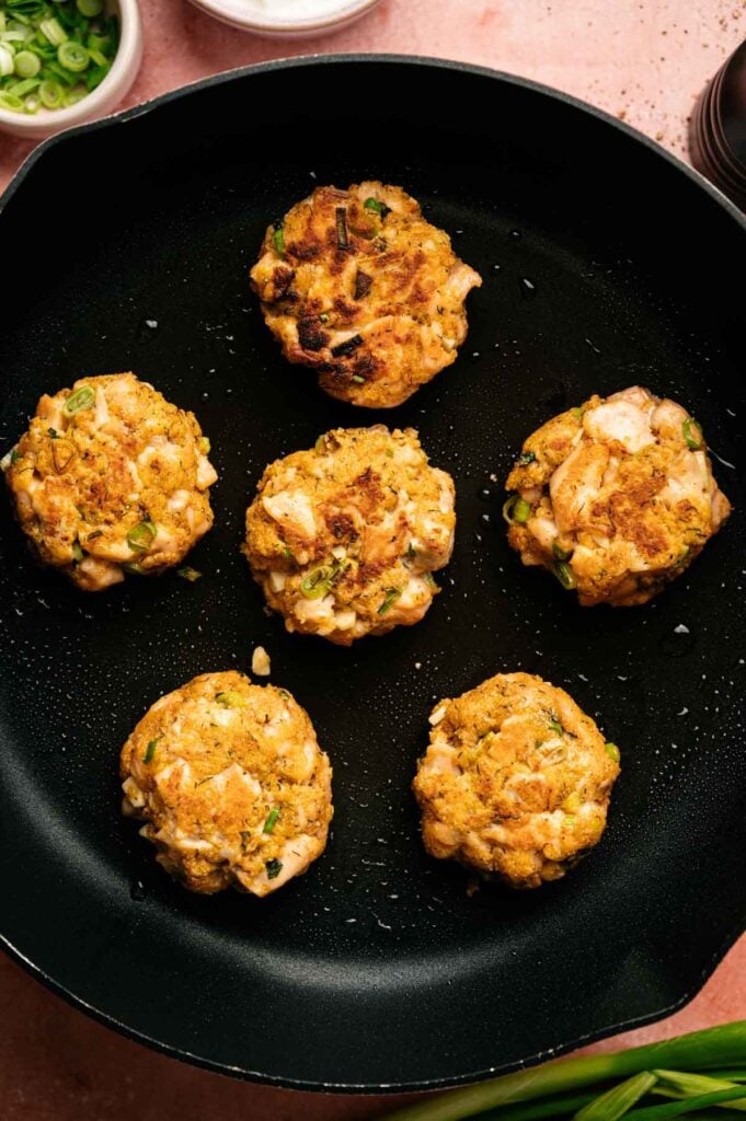 Six golden-brown salmon patties are sizzling in a black skillet, with fresh green herbs visible in the savory mixture.