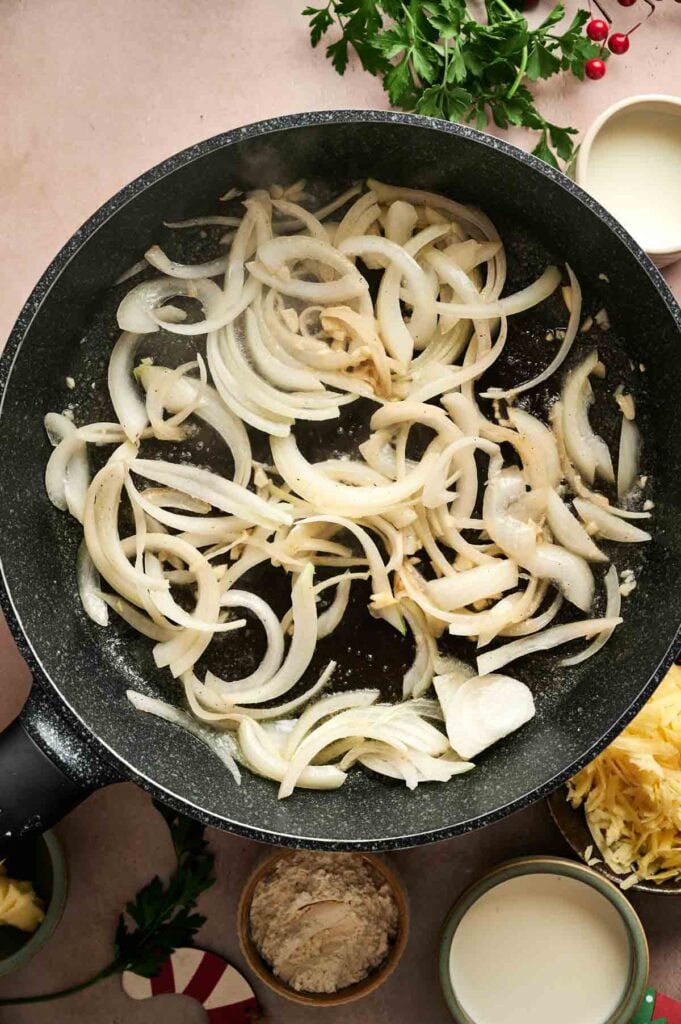 Sliced onions are sautéing in a black pan on a countertop, surrounded by small bowls of ingredients and fresh herbs—setting the perfect stage for preparing comforting dishes like Air Fryer Scalloped Potatoes and Ham.