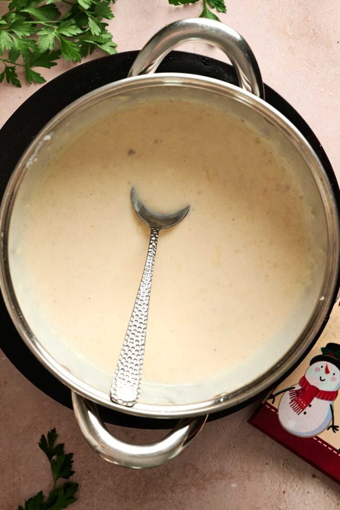 A pot of creamy white soup with a spoon inside, sitting on a light surface, with parsley and a napkin featuring a snowman nearby evokes cozy winter vibes—perfect alongside classic comfort foods like Air Fryer Scalloped Potatoes and Ham.
