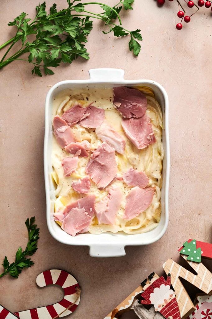 A white baking dish filled with creamy pasta topped with pieces of ham, Air Fryer Scalloped Potatoes and Ham, is surrounded by parsley and holiday-themed decorations on a light surface.