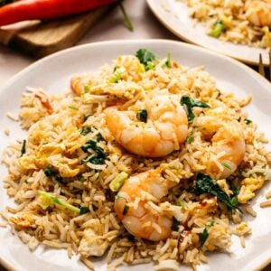 A plate of Thai Fried Rice with shrimp, egg, greens, and herbs, served on a white dish with a chili pepper and parsley in the background.