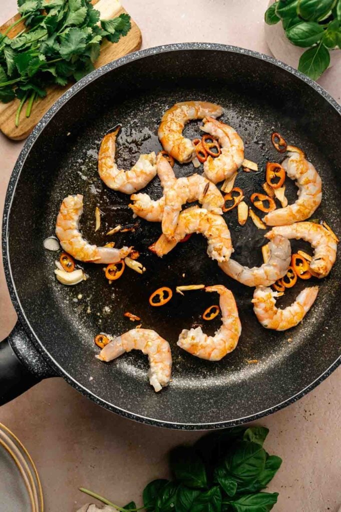 Shrimp and sliced red chili are being sautéed in a black frying pan, hinting at the vibrant flavors of Thai Fried Rice, with some cilantro and basil leaves visible around the pan.