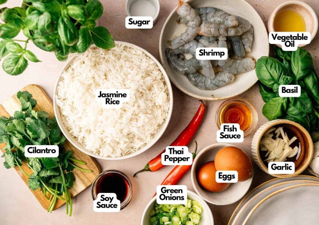 Ingredients for Thai Fried Rice are arranged on a table, including jasmine rice, shrimp, eggs, sauces, vegetables, herbs, chili peppers, and seasonings, each labeled for easy identification.