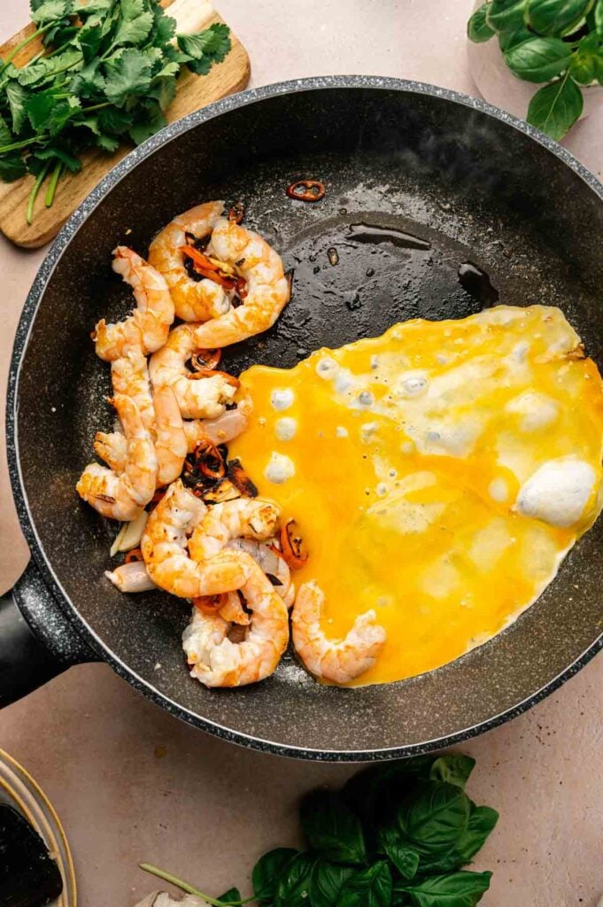 A frying pan with cooked shrimp and partially cooked scrambled eggs, perfect for making Thai Fried Rice, with fresh herbs and a cutting board nearby.