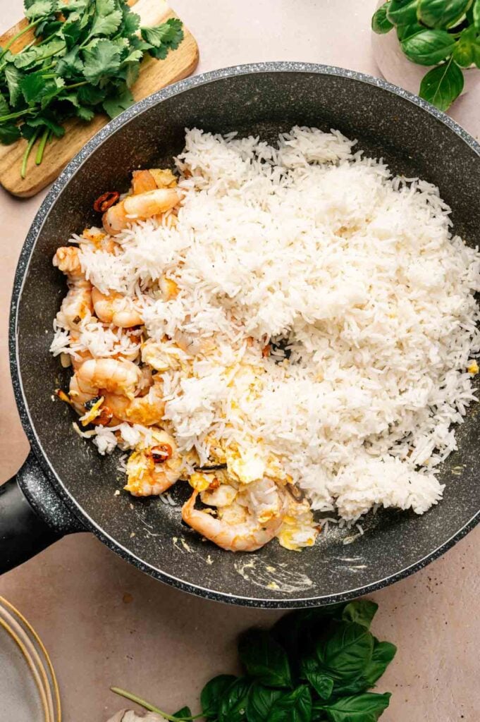 Cooked shrimp, scrambled egg, and white rice in a frying pan, prepared Thai Fried Rice style, with fresh herbs on a cutting board and countertop nearby.