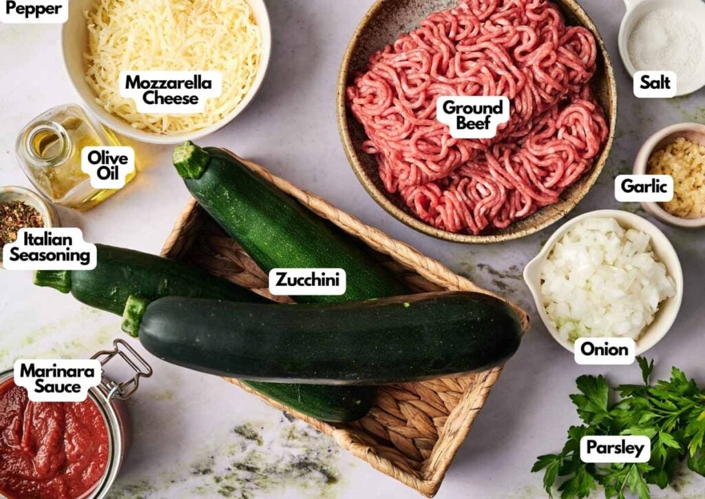 Various labeled ingredients for Air Fryer Zucchini Boats with ground beef and mozzarella cheese are arranged on a countertop, including marinara sauce, onion, garlic, Italian seasoning, and parsley.