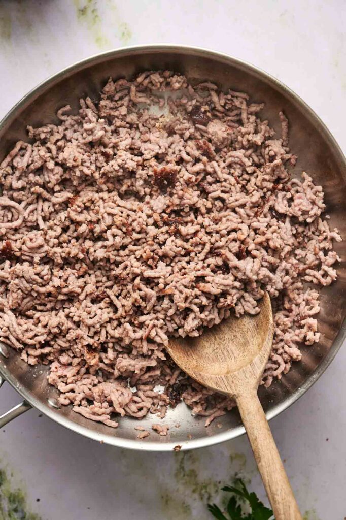 Ground beef being cooked in a stainless steel pan with a wooden spoon stirring the meat, perfect for stuffing into Air Fryer Zucchini Boats.