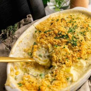 A baking dish filled with creamy Air Fryer Easter Casserole topped with golden breadcrumbs, with a gold spoon scooping some out. Fresh herbs and a pepper grinder are in the background.