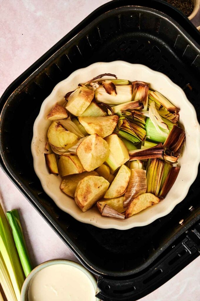A round dish inside an air fryer contains roasted potato chunks and pieces of leek, perfect for starting an Air Fryer Potato and Leek Soup. Raw leeks and a small bowl of sauce are nearby on a pale surface.