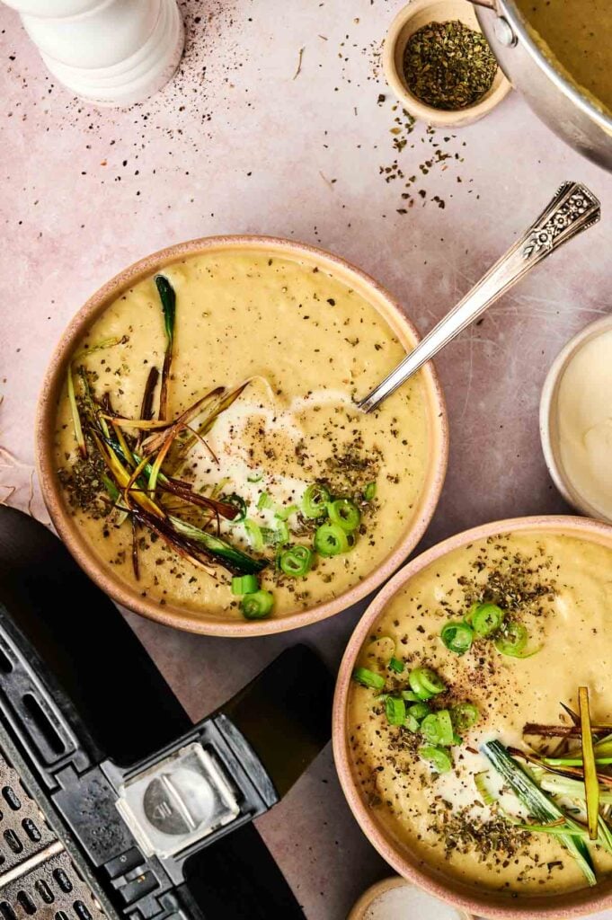 Two bowls of creamy Air Fryer Potato and Leek Soup, garnished with sliced green onions, herbs, and charred leeks, sit on a table next to a pot, a spoon, and a small bowl of dried herbs.
