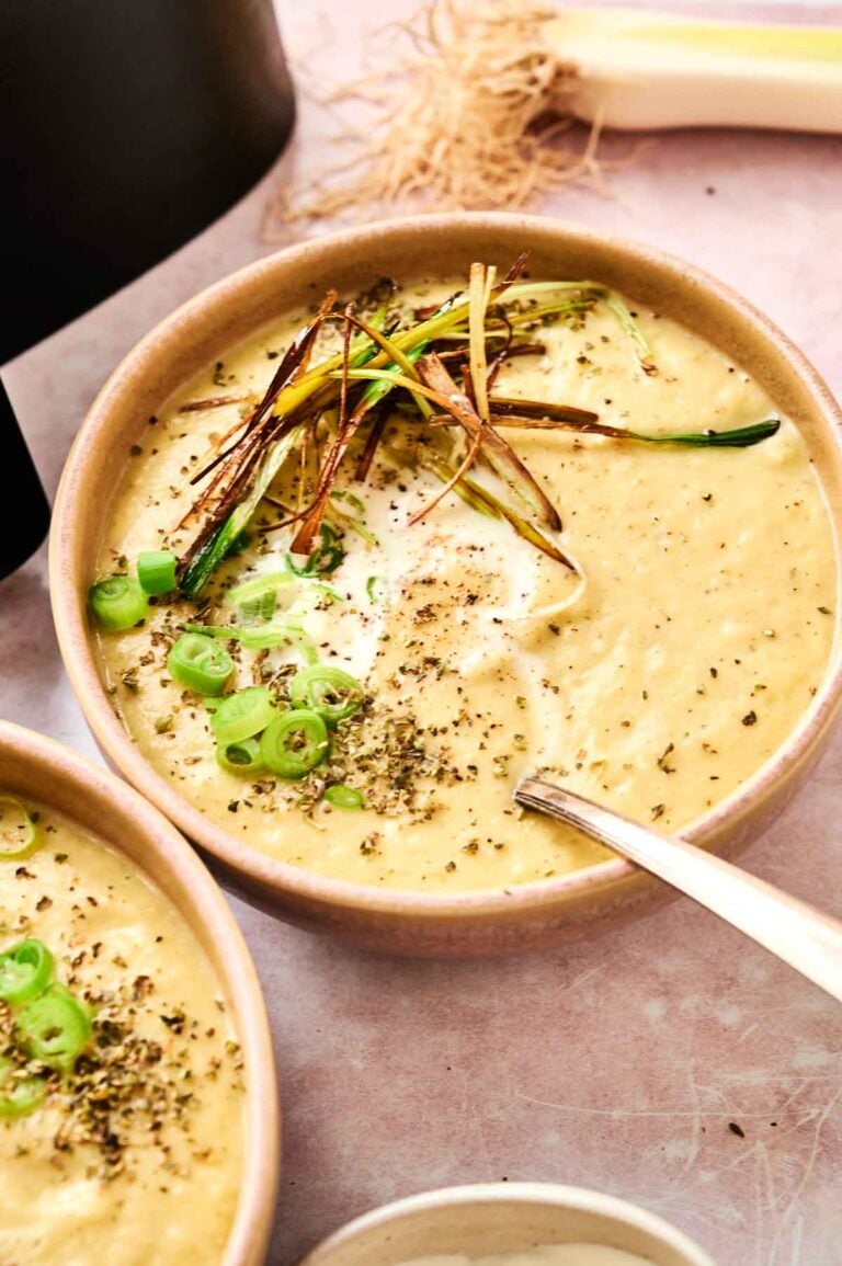 A bowl of creamy Air Fryer Potato and Leek Soup topped with sliced green onions, crispy fried leeks, ground pepper, and herbs, with a spoon resting inside.
