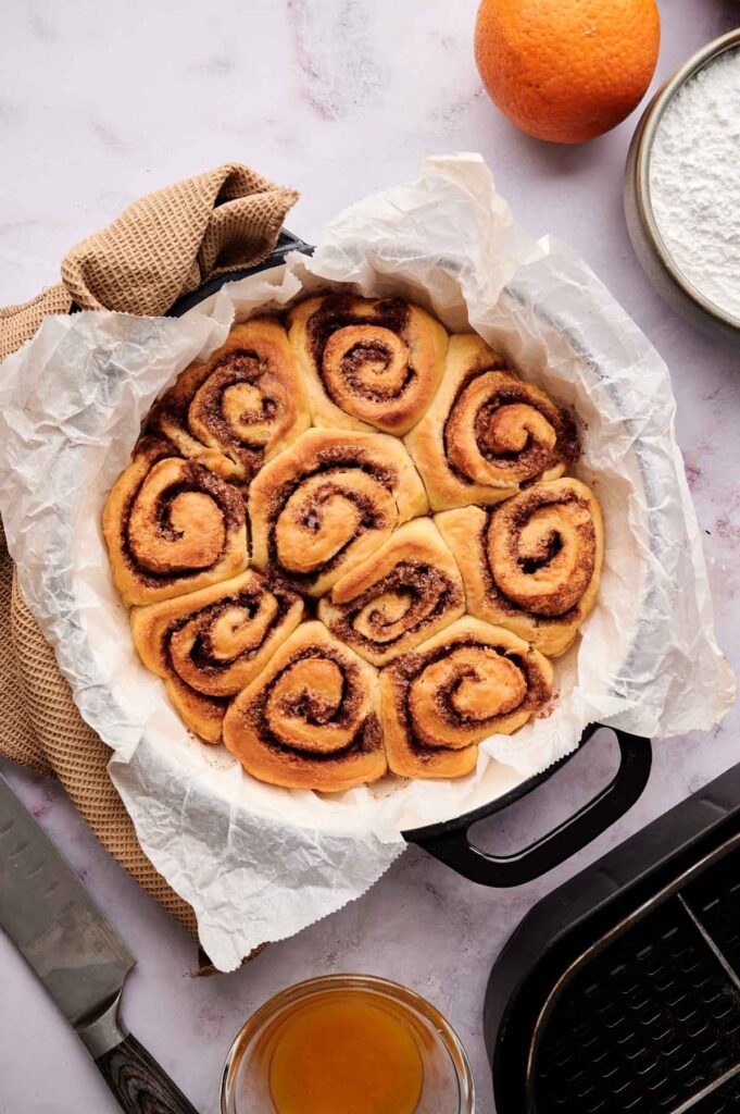 A round pan lined with parchment paper holds nine freshly baked Air Fryer Orange Rolls, with an orange, powdered sugar, a knife, and a bowl of glaze arranged nearby on the countertop.