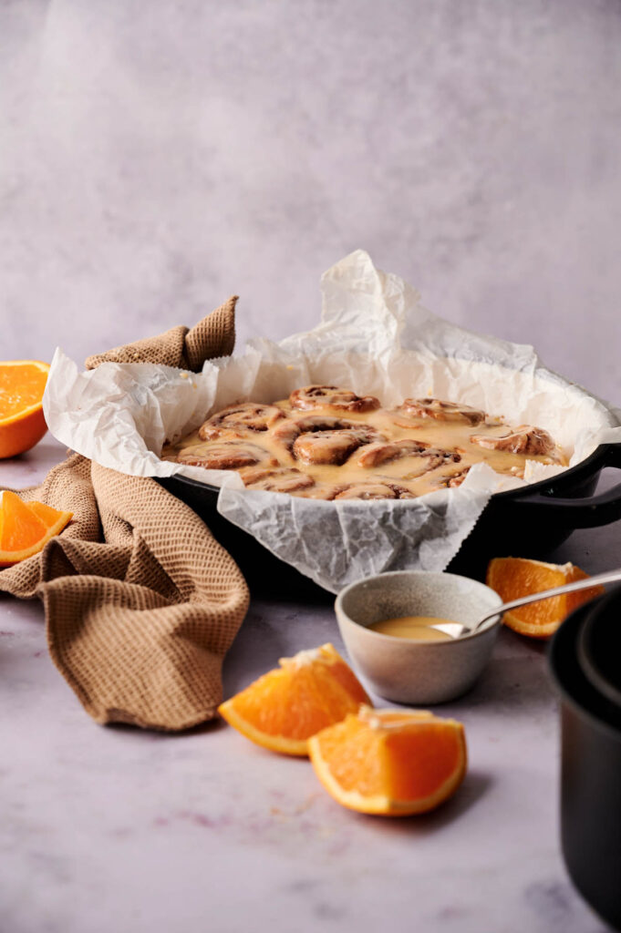 A pan of Air Fryer Orange Rolls on parchment paper, surrounded by orange slices, a bowl of glaze with a spoon, and a brown kitchen towel.