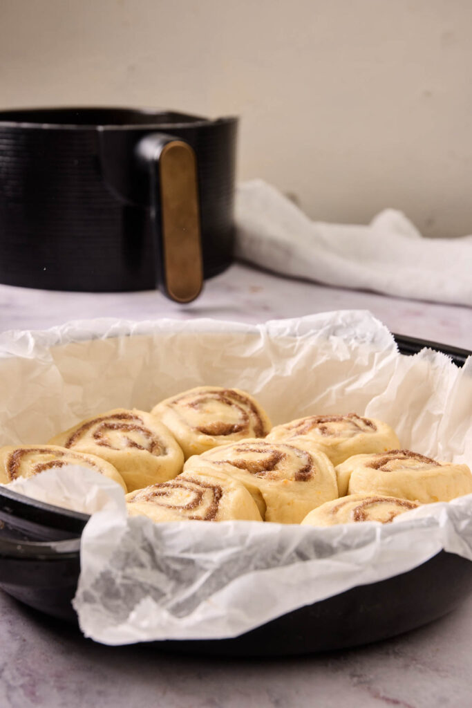 A round black pan lined with parchment paper holds unbaked Air Fryer Orange Rolls. An air fryer is visible in the background.