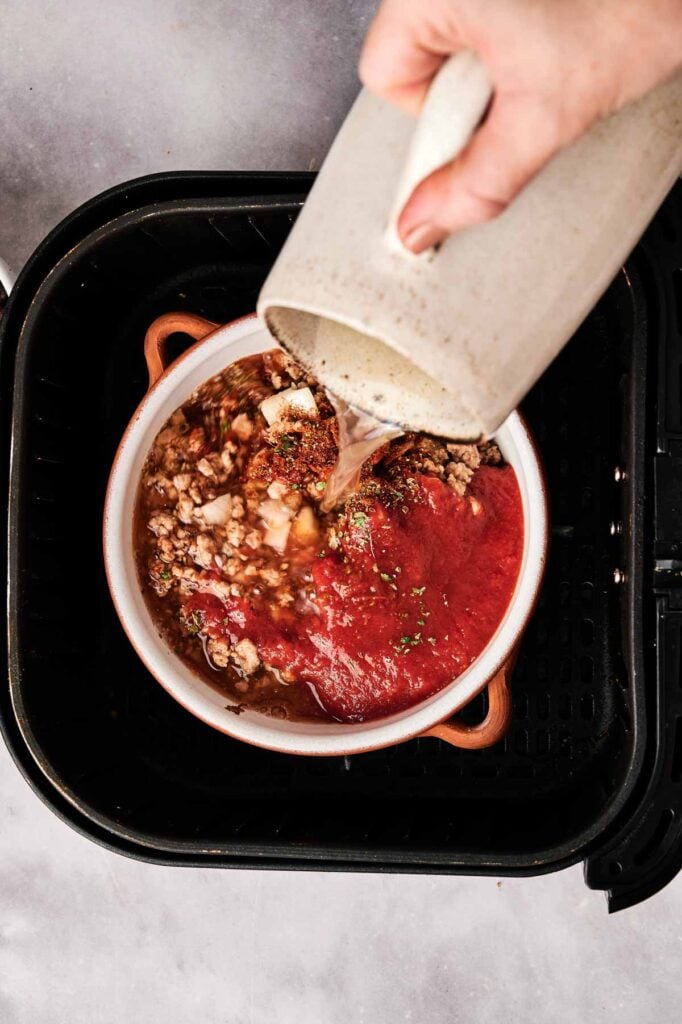 A hand pours liquid from a pitcher into a bowl of ground meat, tomato sauce, and seasonings, preparing Air Fryer Taco Soup inside an air fryer.