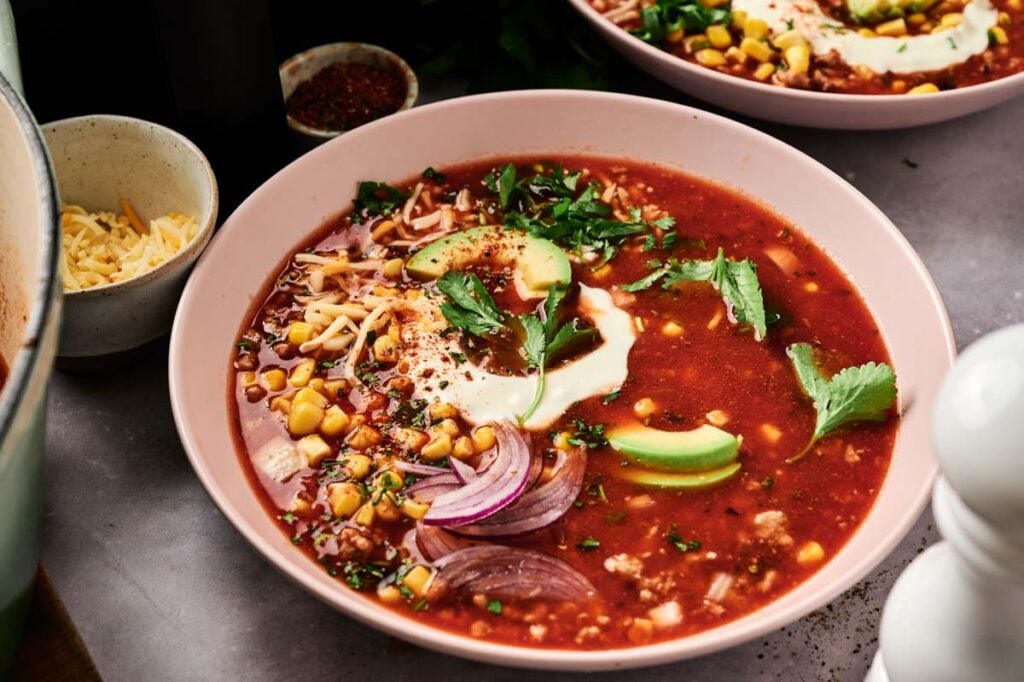 A bowl of Air Fryer Taco Soup topped with avocado slices, red onion, corn, shredded cheese, cilantro, and a dollop of sour cream.
