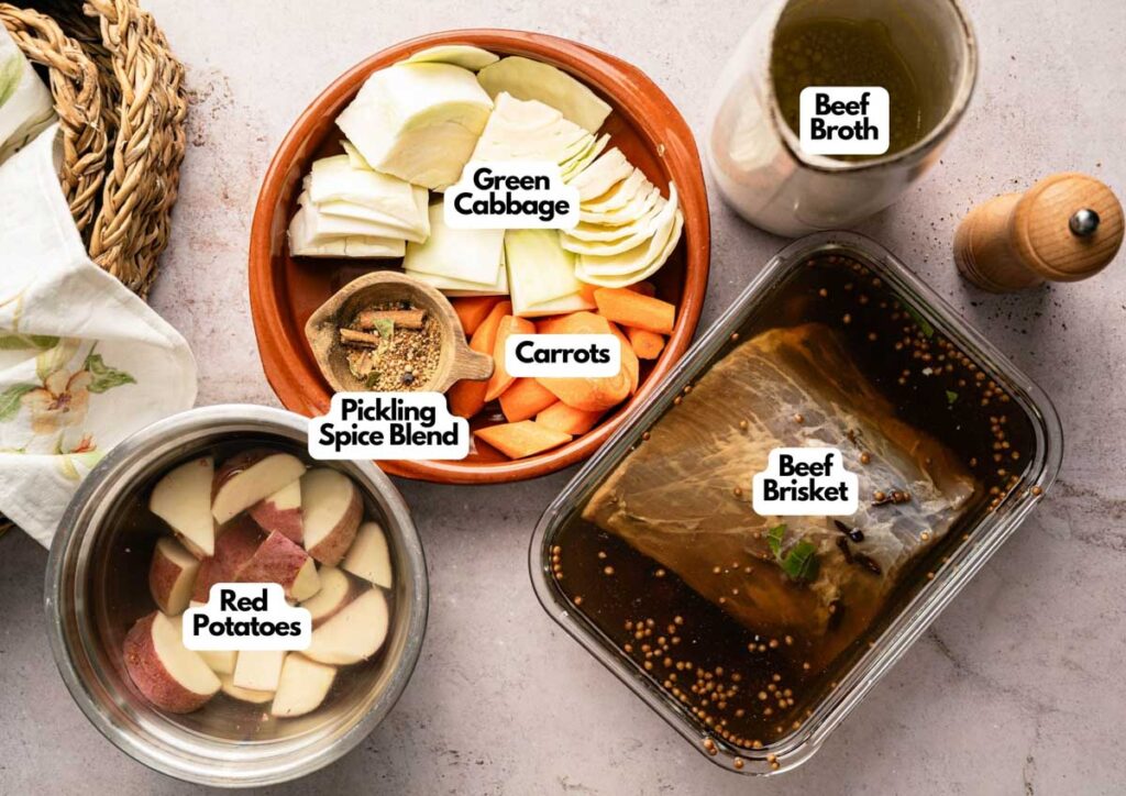 Labeled ingredients for Corned Beef and Cabbage—green cabbage, red potatoes, carrots, pickling spice blend, beef brisket in marinade, and beef broth—are neatly arranged on a countertop.