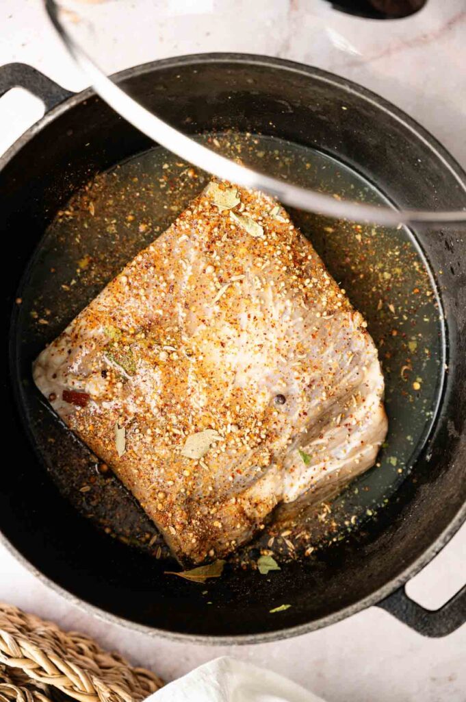 A seasoned piece of raw meat, perfect for Corned Beef and Cabbage, sits in a black pot with liquid, ready for cooking.