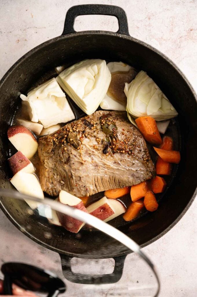 A pot contains a flavorful Corned Beef and Cabbage brisket surrounded by chopped cabbage, potatoes, and carrots, with some savory broth visible at the bottom.