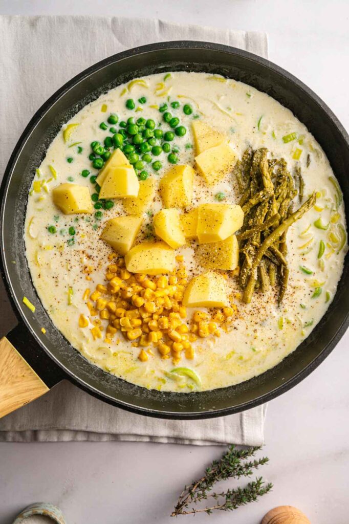 A skillet filled with a creamy white sauce, uncooked potato chunks, green peas, corn, and green beans, seasoned with black pepper—perfect for an Easter Casserole that’s both hearty and festive.