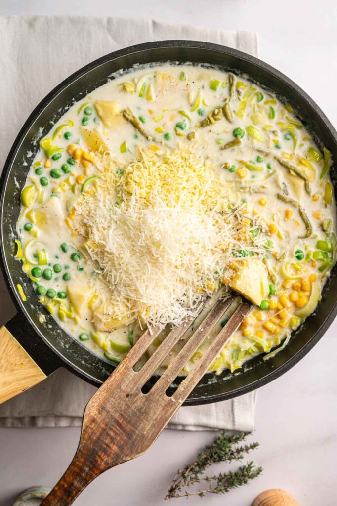 A skillet filled with creamy vegetable pasta, reminiscent of an Easter Casserole, is topped with grated cheese and sits on a light napkin with a wooden spatula resting inside.