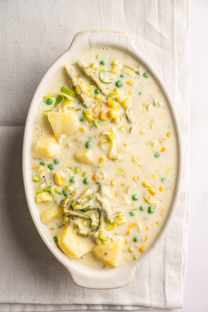 Oval dish filled with creamy vegetable chowder containing potatoes, peas, corn, and leafy greens, reminiscent of a comforting Easter Casserole, placed on a light-colored cloth.
