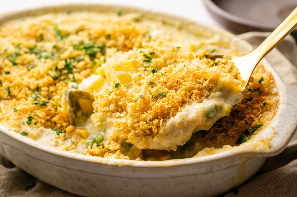 A serving spoon lifts a portion of creamy Easter Casserole with a golden breadcrumb topping from a white baking dish.