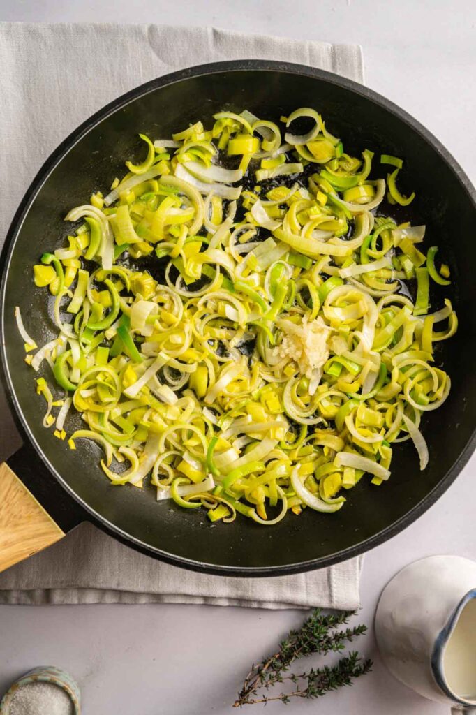Sliced leeks sautéing in a black frying pan on a light cloth, with sprigs of thyme and a salt container nearby—an inviting base for your next Easter Casserole.