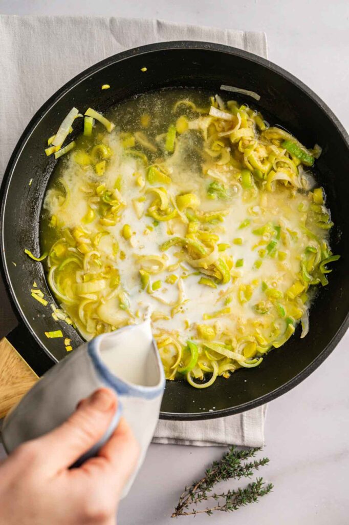 A hand pours milk into a skillet containing chopped leeks cooking in liquid, the creamy mixture forming the perfect base for an Easter Casserole. Fresh thyme sprigs and a gray napkin are nearby.