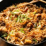 A pan of yakisoba stir-fried noodles with chicken, vegetables, green onions, and savory sauce, with chopsticks resting on the edge.