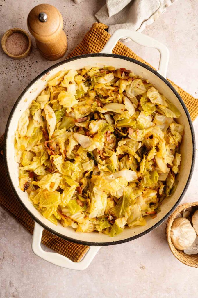 A white pot of fried cabbage and bacon sits on a brown mat, surrounded by a pepper grinder, a small bowl of salt, and whole garlic bulbs.