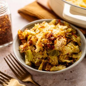 A bowl of fried cabbage topped with red pepper flakes sits beside two forks and a jar of spices on a light surface.