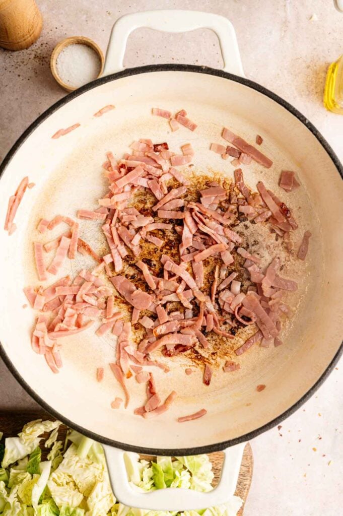 A white pot contains cooking strips of ham, some browned, with a bit of oil. Chopped cabbage, ready to be added for delicious fried cabbage, is visible in the corner. Salt and oil containers are nearby.