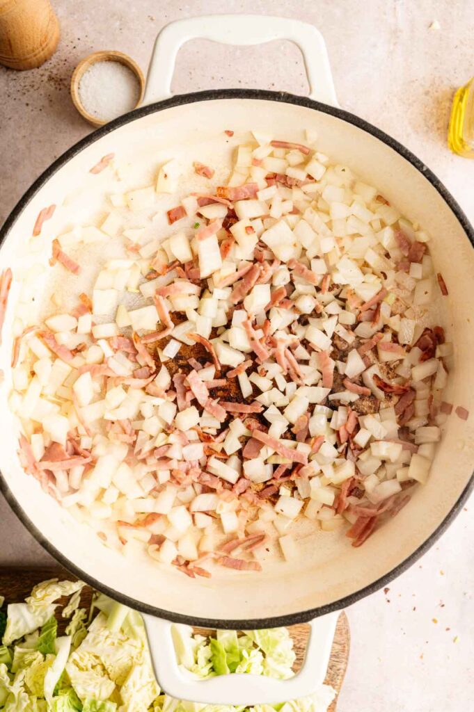 Chopped onions and bacon are sautéing in a white pot, with a bowl of salt, a bottle of oil, and chopped cabbage nearby—perfectly setting the stage to make delicious Fried Cabbage.