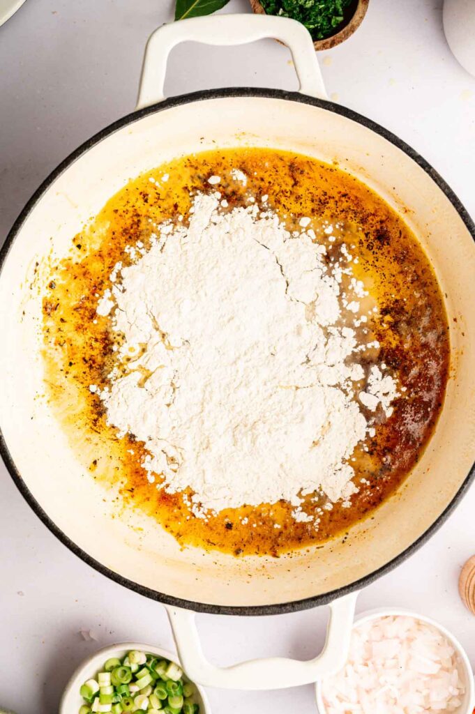 A white pot with browned oil and seasonings, perfect for starting a flavorful gumbo, is topped with a mound of flour and sits on a white surface surrounded by small bowls of chopped green onions and other ingredients.