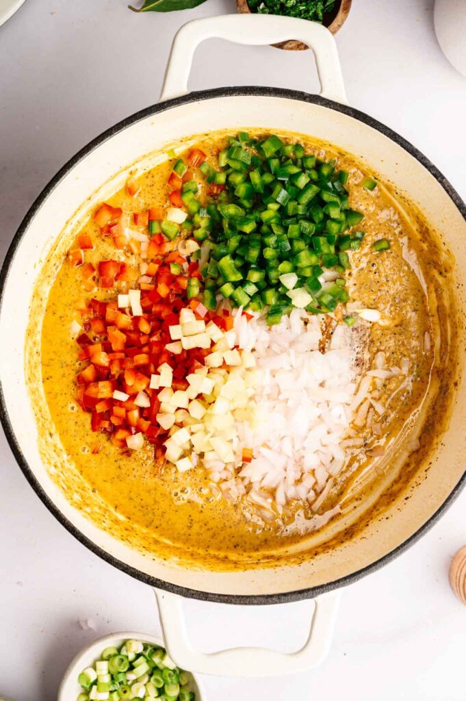 A pot with diced red bell pepper, green bell pepper, onion, and celery—the classic gumbo trinity—arranged on top of a yellow seasoned mixture, ready to be stirred.