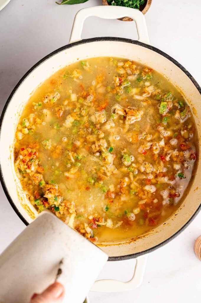 A person pours liquid into a white pot filled with a chunky gumbo mixture of vegetables and broth on a white surface.