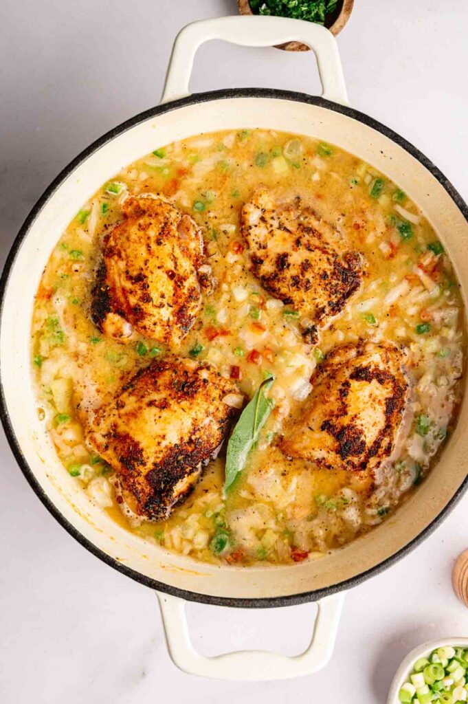 Four seasoned chicken thighs simmer in a pot with a creamy, gumbo-inspired, vegetable-filled sauce and a bay leaf, ready for cooking.