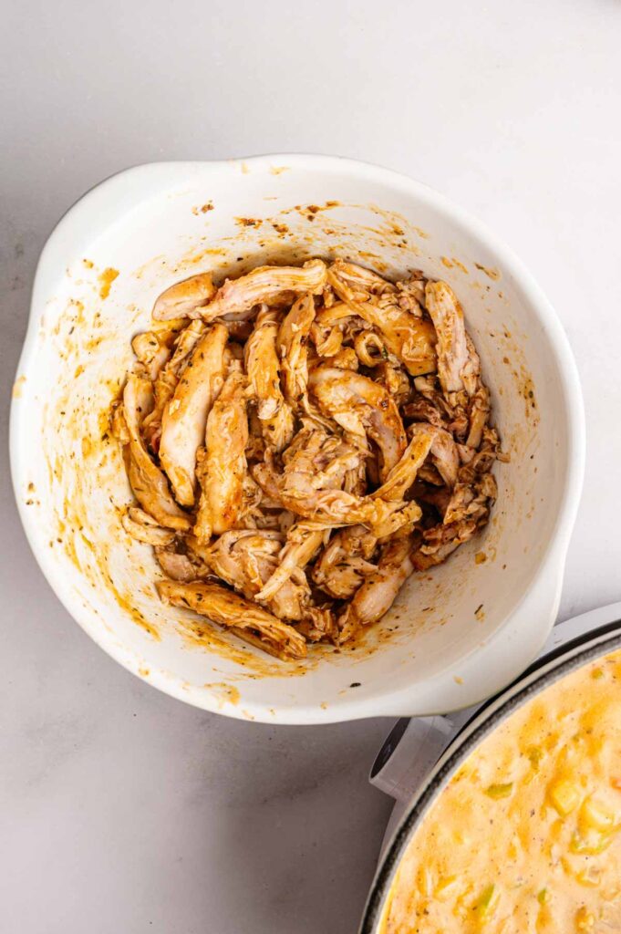 Shredded, seasoned chicken in a white mixing bowl, with a partial view of a creamy gumbo dish in a pot nearby on a light surface.