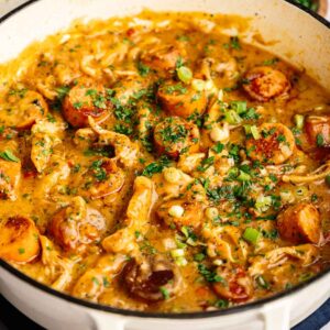 A white pot filled with savory gumbo, brimming with sausage, chicken, and vegetables, garnished with chopped green onions and parsley.