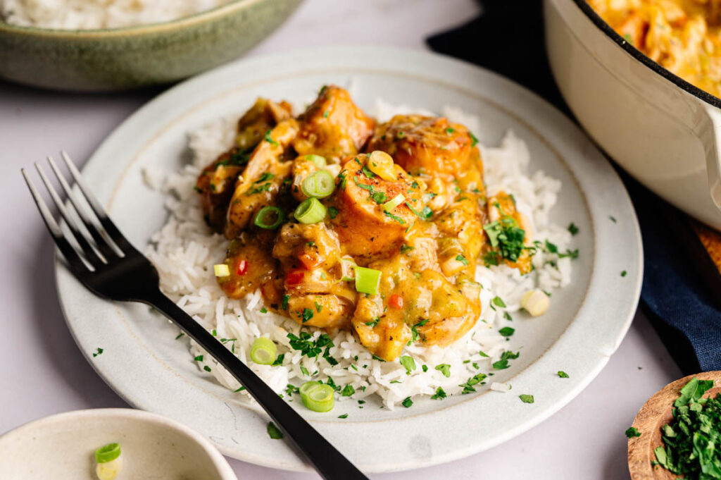 A plate of white rice topped with creamy chicken curry, garnished with chopped green onions and herbs, reminiscent of gumbo flavors, with a black fork on the side.