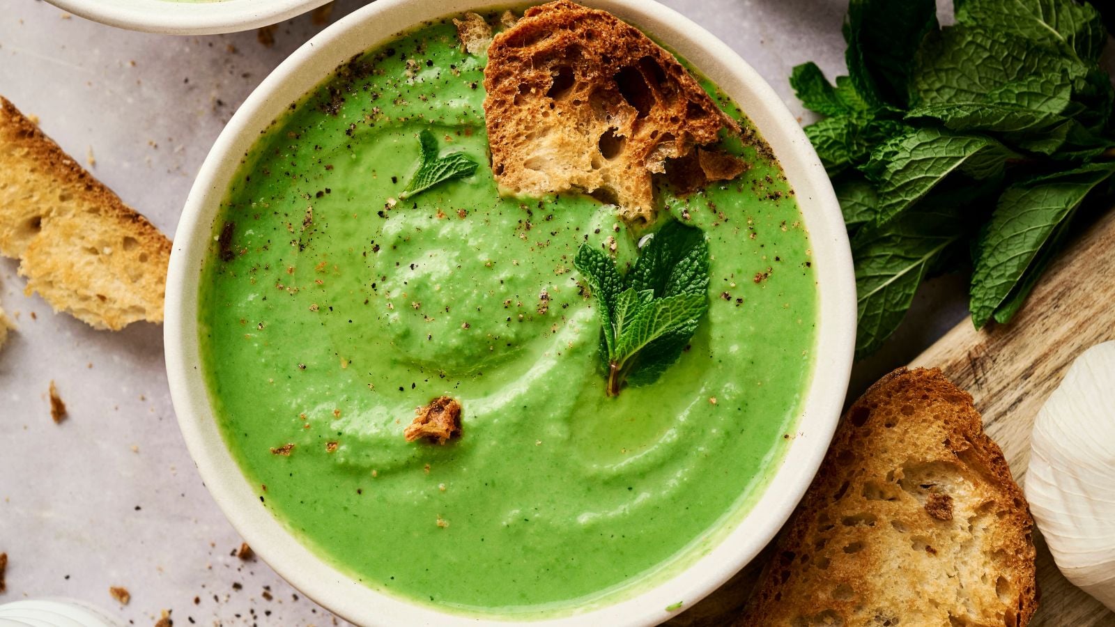 A bowl of green soup garnished with mint leaves and croutons, with slices of bread and fresh mint on the side.
