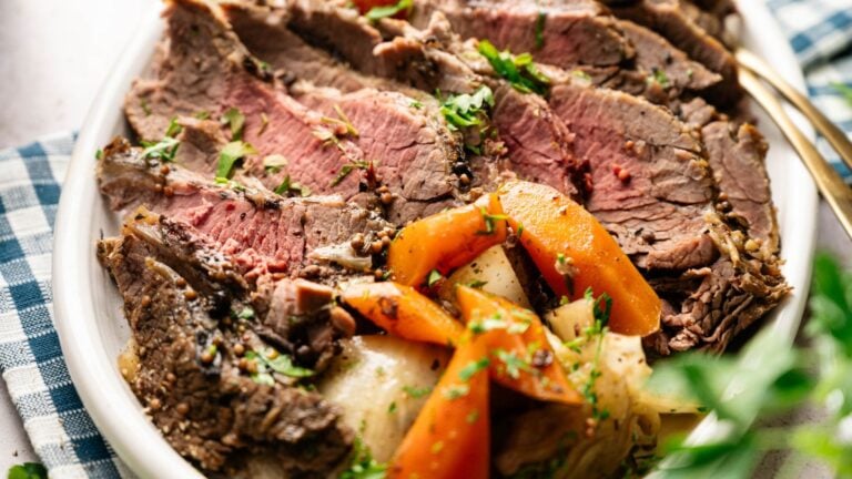 Sliced roast beef served on a plate with cooked carrots and potatoes, garnished with chopped herbs.