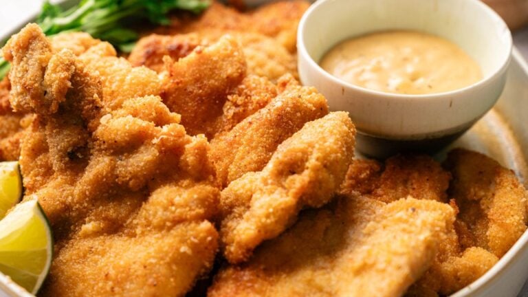 Close-up of crispy breaded chicken cutlets with lime wedges and a small bowl of dipping sauce on the side.