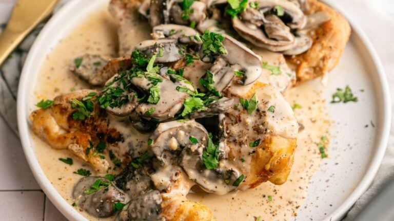 A plate of cooked chicken breast topped with creamy mushroom sauce and garnished with chopped parsley.