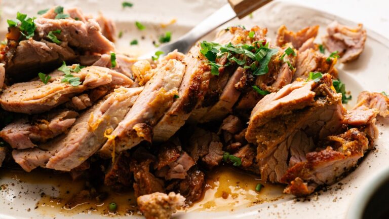 Slices of cooked, seasoned pork garnished with chopped herbs are arranged on a plate, with a fork resting nearby.